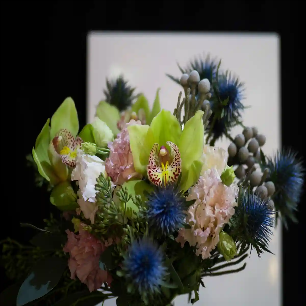 Bouquet of flowers with green orchids, blue thistles, and pink roses against a blurred background.