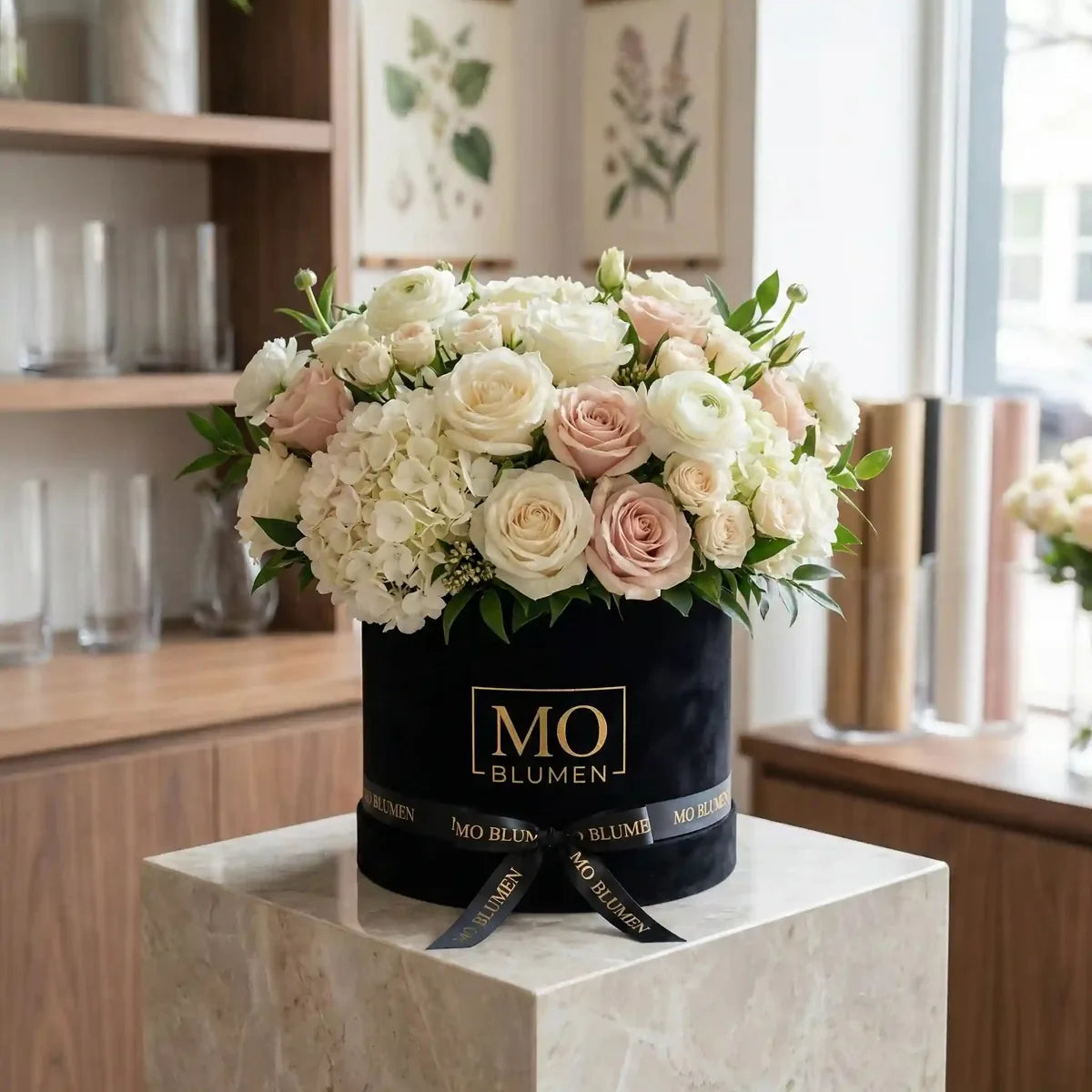 Schwarze Samt-Blumenbox mit hellrosa und weißen Rosen, Ranunkeln und Hortensien von MO BLUMEN, mit goldenem Logo und schwarzem Samtband, auf einem Marmorsockel.