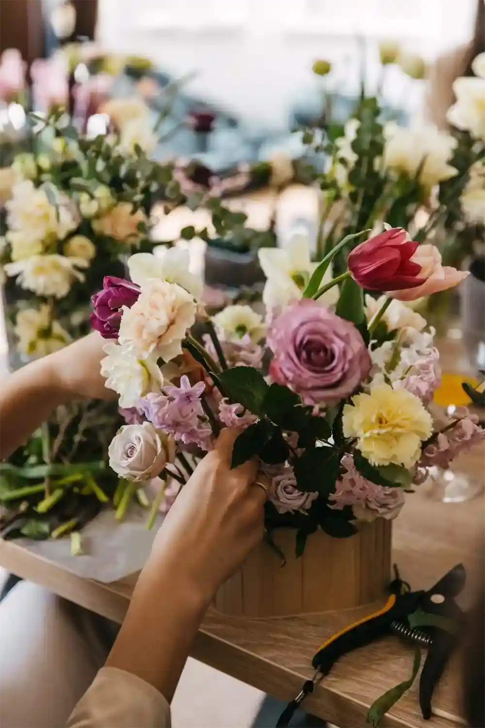 Person arranging flowers in a wooden basket with a blurred floral background