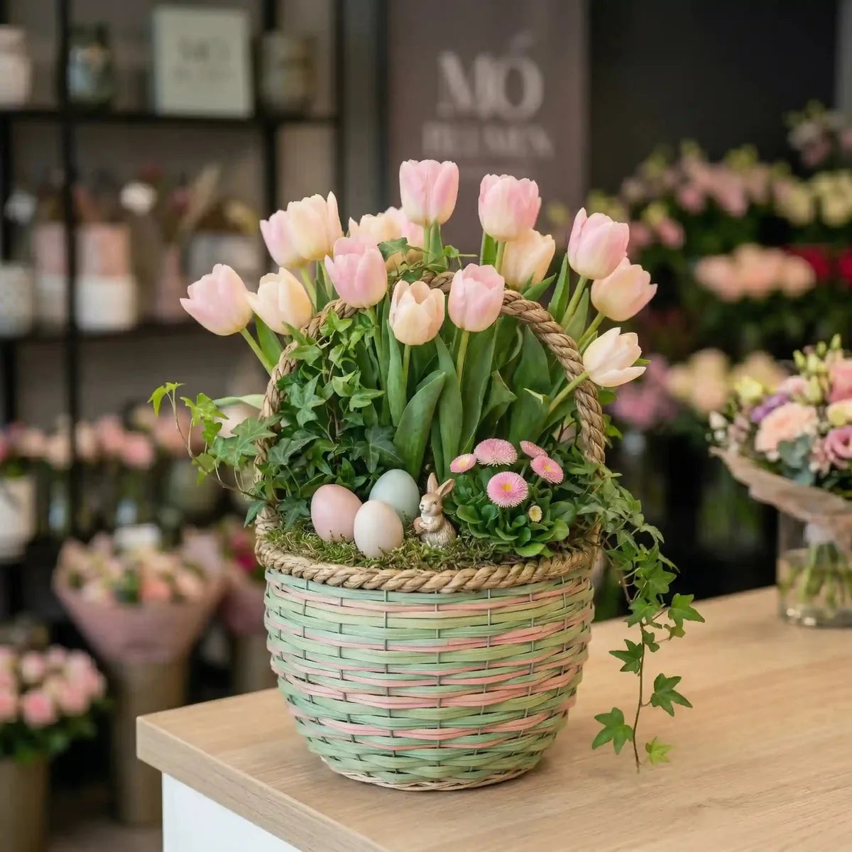 Zarter Frühlingskorb mit rosa-cremefarbenen Tulpen von MO BLUMEN, geflochten in Hellgrün-Rosa, mit Ostereiern, Hase und Efeu auf einem Holztisch.
