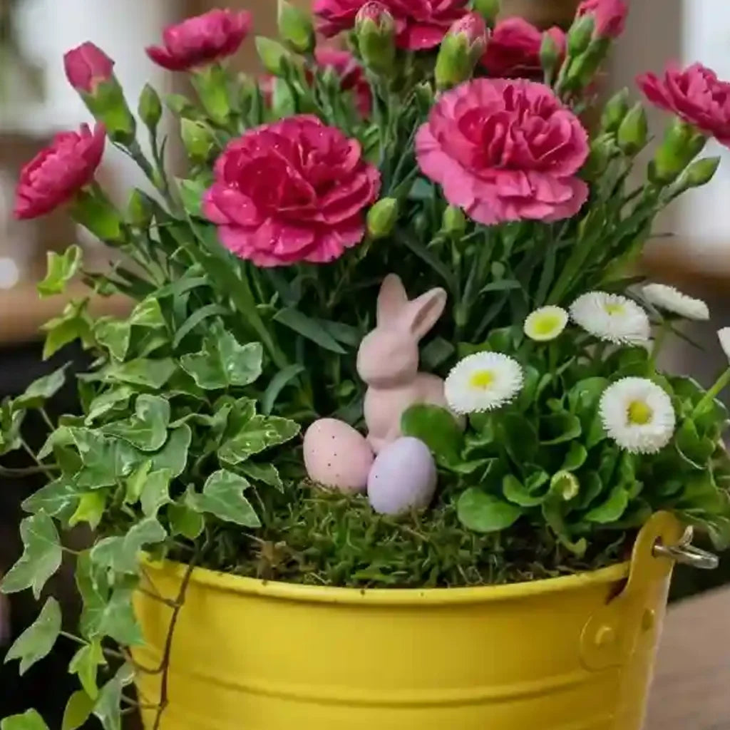 Frühlingskorb mit rosa Nelken und Margeriten von MO BLUMEN in einem gelben Eimer, mit rosa Hase und Ostereiern auf Moos.
