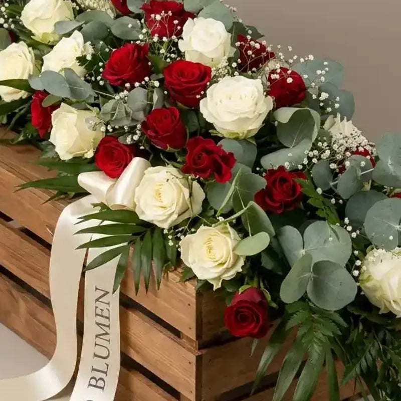 Sargbukett aus rot-weißen Rosen und Eukalyptus von MO BLUMEN, auf einer Holzkiste mit weißem Schleifenband, beschriftet mit BLUMEN.