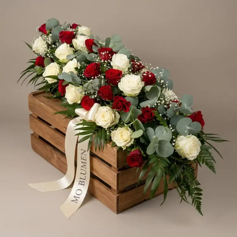 Sargbukett aus rot-weißen Rosen, Eukalyptus und Schleierkraut von MO BLUMEN, auf einer Holzkiste mit beschriftetem Band.