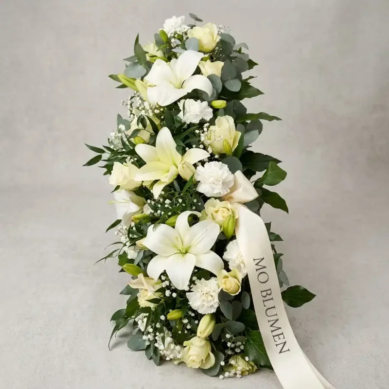 Ein Sargbukett mit weißen Lilien, Nelken und Rosen von MO BLUMEN, dekoriert mit Grünpflanzen und einem cremefarbenen Schleifenband.