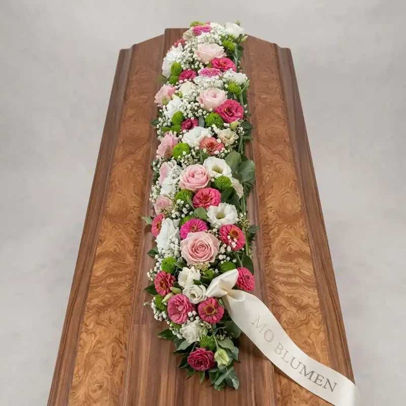 Sargschmuck mit rosa Lisianthus, weißen und grünen Blumen von MO BLUMEN auf einem gemaserten Holzsarg mit weißer Schleife.