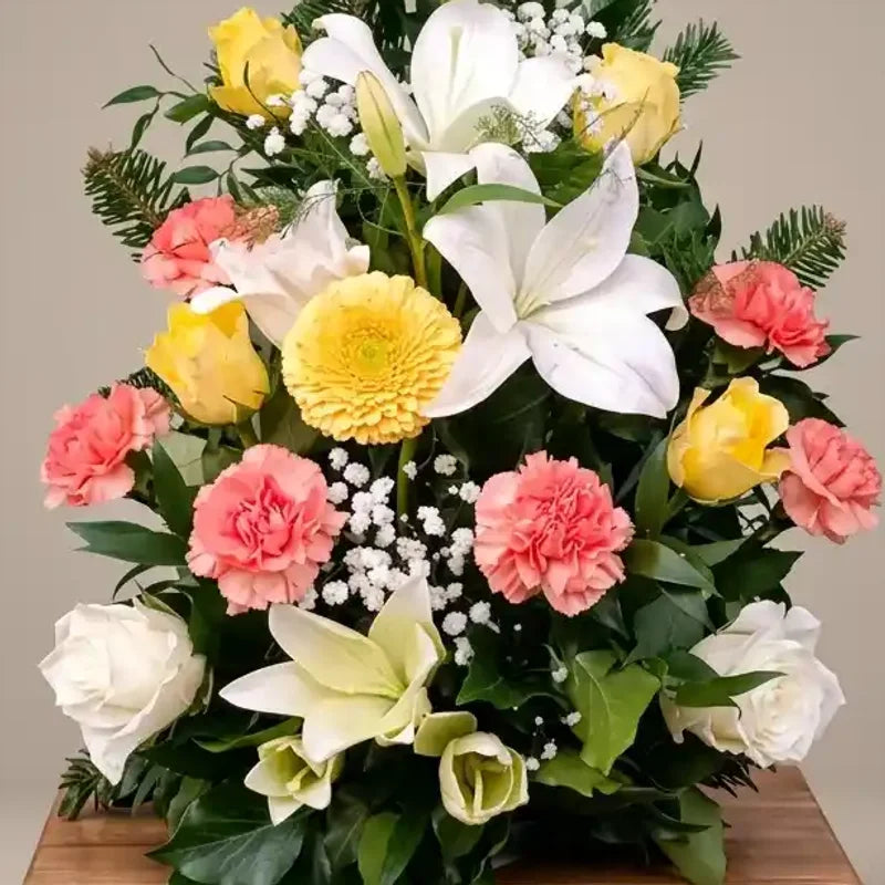 Aufrechtes buntes Trauergesteck von MO BLUMEN mit weißen Lilien, gelben und weißen Rosen, rosa Nelken, gelber Gerbera und grünen Zweigen.