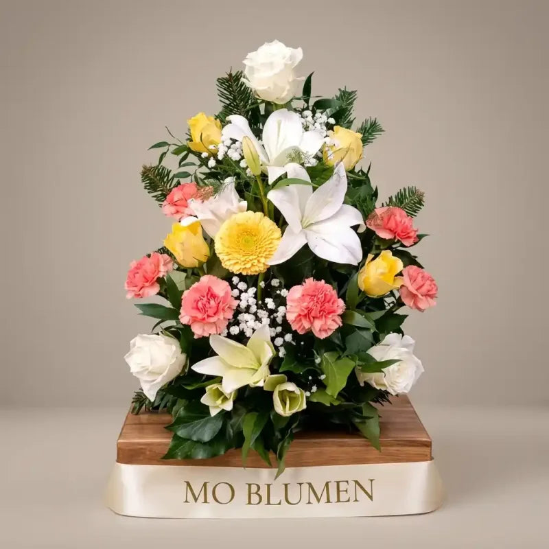 Ein aufrechtes, buntes Trauergesteck von MO BLUMEN mit weißen Rosen, gelben Lilien, pinkfarbenen Nelken und gelben Gerbera auf einem Holzsockel mit cremefarbenem Band.