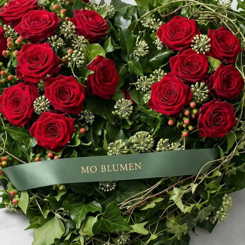 Herzförmiges Trauergesteck mit roten Rosen von MO BLUMEN, umgeben von grünem Efeu und Beeren mit einem grünen Band.