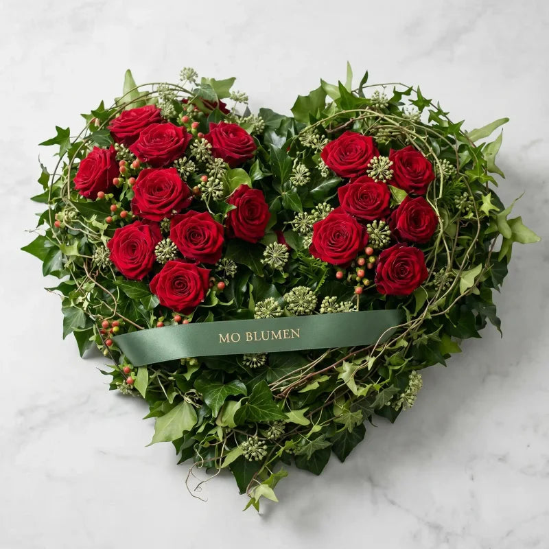 Herzförmiges Trauergesteck mit roten Rosen von MO BLUMEN, dekoriert mit Efeu, kleinen roten Beeren und einem grünen Band.