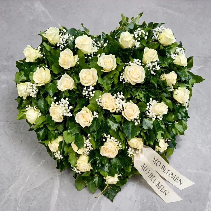 Herzförmiges Trauergesteck mit weißen Rosen von MO BLUMEN, dekoriert mit Efeublättern, Schleierkraut und zwei Bändern mit dem Schriftzug MO BLUMEN.
