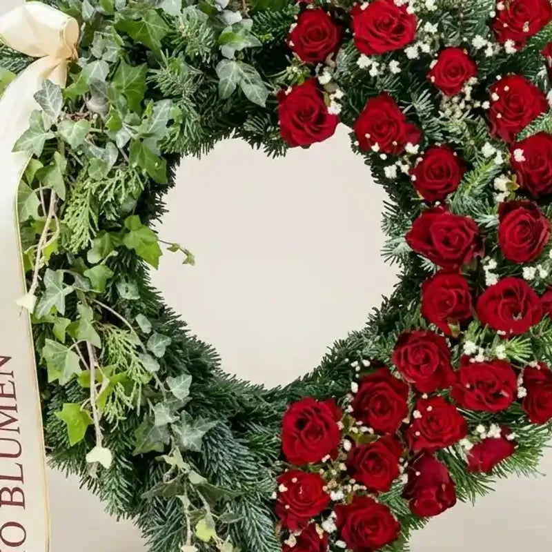 Trauerherz aus Efeu und roten Rosen von MO BLUMEN, dekoriert mit Tanne und weißem Schleifenband.