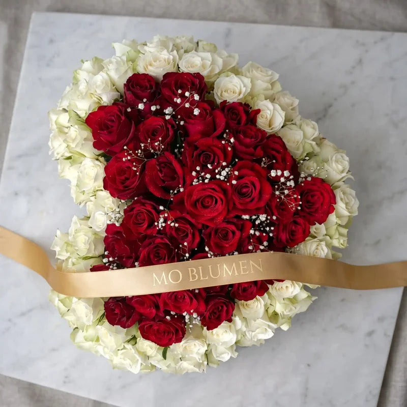 Trauerherz aus rot-weißen Rosen von MO BLUMEN, mit kleinen weißen Blüten und goldenem Band auf Marmor.