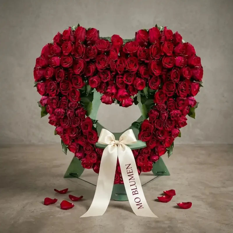 Luxuriöses stehendes Trauerherz aus roten Rosen von MO BLUMEN mit gebrandetem cremefarbenen Band und verstreuten Blütenblättern.