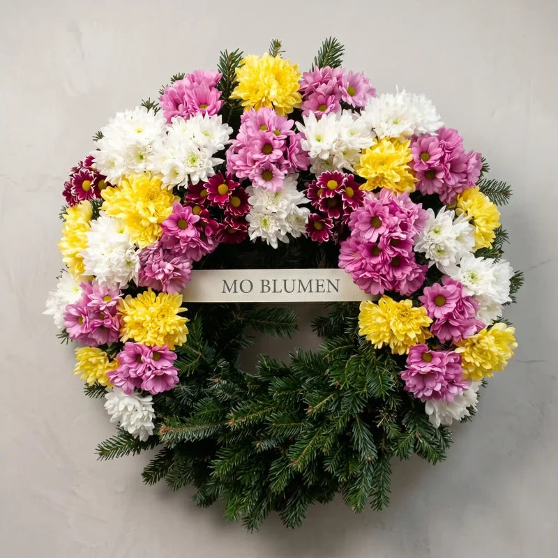 Bunter Trauerkranz aus Chrysanthemen von MO BLUMEN auf Tannengrün, mit weißen, gelben, rosa und dunkelroten Blüten und einem Textband.