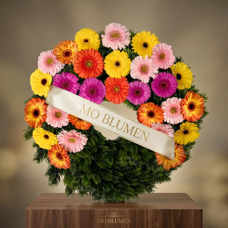 Regenbogenfarbener Gerbera-Trauerkranz von MO BLUMEN, mit weißem Band mit goldenem MO BLUMEN-Schriftzug, auf einem Holzpodest.