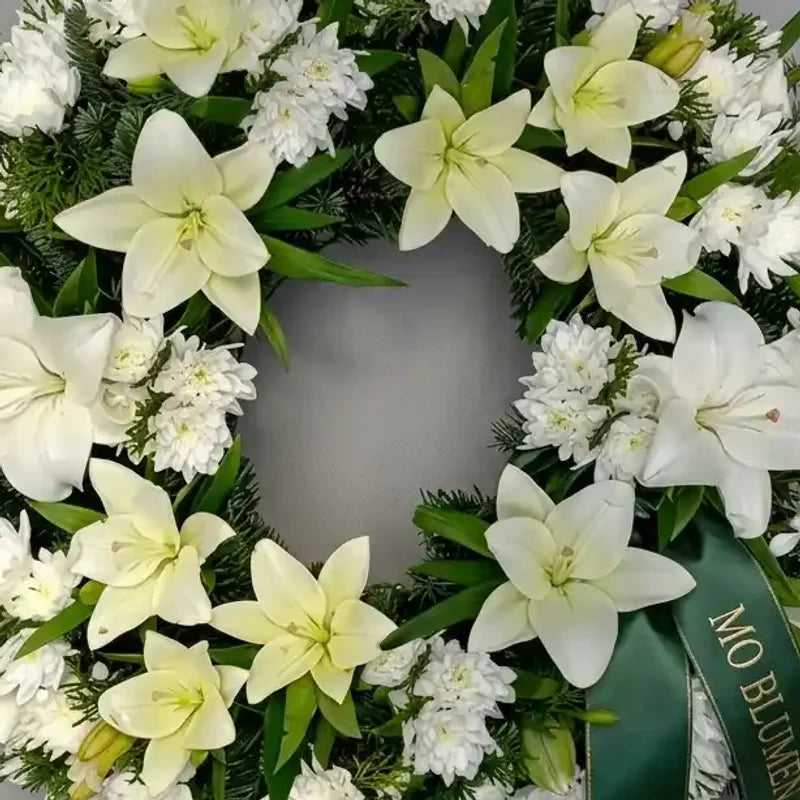 Trauerkranz mit weißen Lilien und Nelken von MO BLUMEN, dekoriert mit grünen Zweigen und einem grünen Band.
