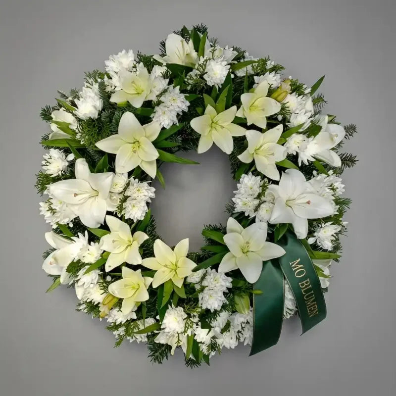 Weißer Trauerkranz aus Lilien und Nelken von MO BLUMEN, reich geschmückt mit Grün und grünem Schleifenband mit goldener Aufschrift.
