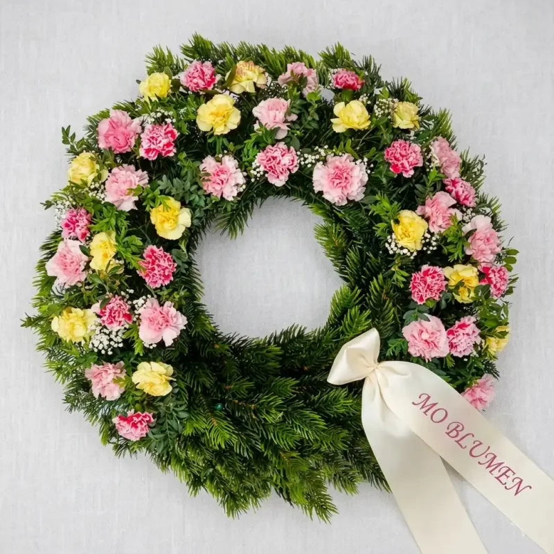 Runder Trauerkranz aus Tanne mit rosa und gelben Nelken von MO BLUMEN, verziert mit cremefarbenem Band und Schriftzug 'MO BLUMEN'.
