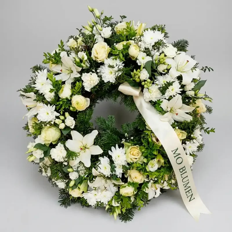 Trauerkranz in reinem Weiß von MO BLUMEN mit weißen und hellgelben Blumen, grünem Beiwerk und cremefarbenem Band mit Aufschrift 'MO BLUMEN'.
