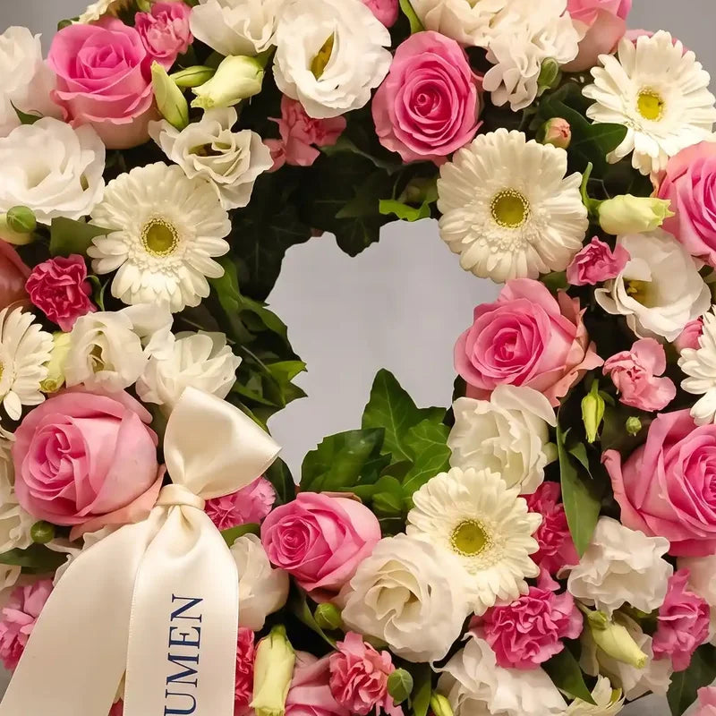 Trauerkranz in Rosa und Weiß von MO BLUMEN, mit Rosen, Gerbera, Lisianthus, grünen Blättern und weißem Band mit Text 'UMEN'.