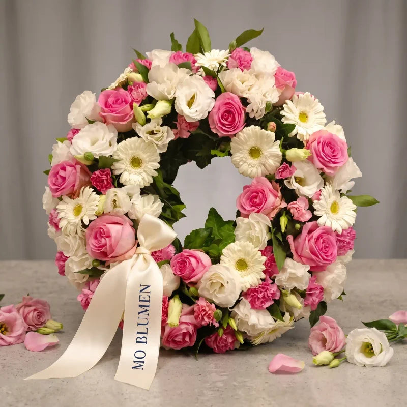Rosa-weißer Trauerkranz von MO BLUMEN mit cremefarbener Schleife, bedruckt mit MO BLUMEN, und zarten Blüten.
