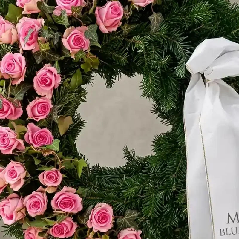 Trauerkranz aus Tannengrün mit rosa Rosen von MO BLUMEN, dekoriert mit Efeublättern und einer weißen Schleife mit goldener Kante.