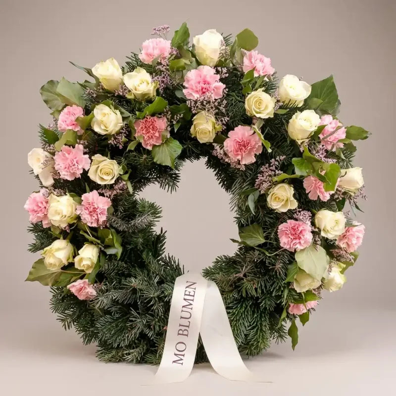 Trauerkranz aus Rosen und Nelken in Rosé von MO BLUMEN mit weißem Band und grüner Tannengrundlage.