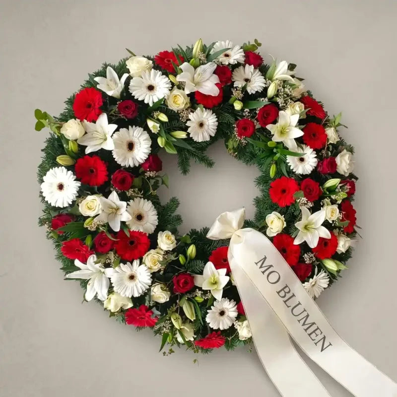 Rot-weiß gemischter Trauerkranz von MO BLUMEN aus Lilien, Rosen und Gerbera auf Tannengrün mit beschrifteter Schleife.