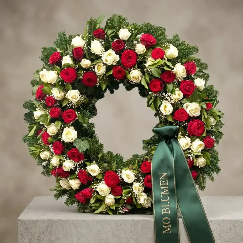 Luxuriöser Trauerkranz mit roten und weißen Rosen von MO BLUMEN, dekoriert mit Tannengrün, kleinen weißen Blüten und einem grünen Schleifenband.