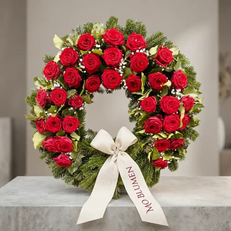 Klassischer Trauerkranz mit roten Rosen von MO BLUMEN, aus Tannengrün, mit einem beige-weißen Band, das "MO BLUMEN" zeigt.