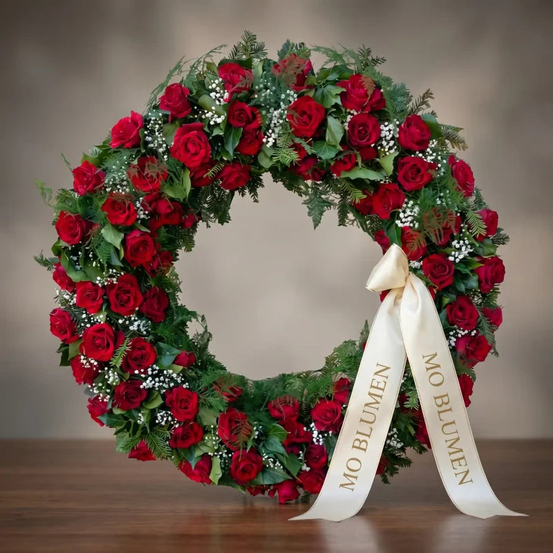 Luxuriöser Vollkranz aus roten Rosen von MO BLUMEN, mit grünem Beiwerk, kleinen weißen Blüten und beigem Schleifenband mit MO BLUMEN Schriftzug.